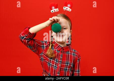 Funny felice bambina con palla di natale decorazione. Un bambino in un abito rosso e manopole con archetto su uno sfondo colorato. Foto Stock