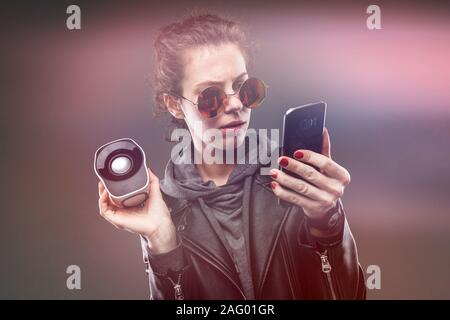 Ragazza caucasica in bicchieri con un altoparlante portatile e un telefono su uno sfondo nero con effetto di alone. Foto Stock