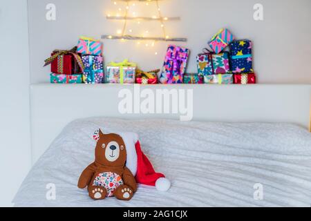 Accogliente orso giocattolo in santa hat sul letto su sfondo colorato e confezioni regalo e albero di Natale fatto di bastoni di legno con una ghirlanda sulla parete. Semplice, Foto Stock