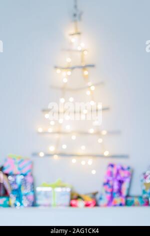 Sfocare lo sfondo di un insolito albero di Natale fatto di bastoni di legno con luci festose sulla parete bianca e colorata confezioni regalo. Semplice, conscie minima Foto Stock