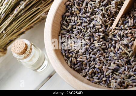 Lavanda essiccata le erbe in un recipiente di legno con convogliatore accanto a olio essenziale bottiglia con sughero e mazzo di fiori di steli in background. Foto Stock