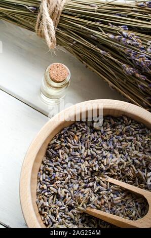 Un mazzetto legato di essiccato Lavanda fiori accanto a una ciotola di legno e scoop contenente le gemme e una bottiglia tappato di olio essenziale. Medicinali a base di erbe e aromaterapia c Foto Stock