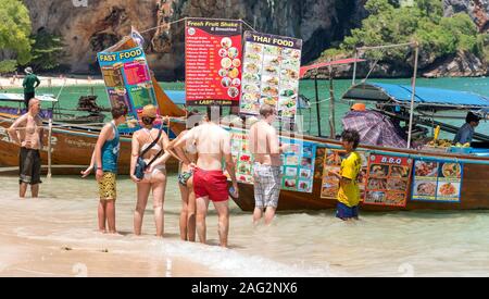 Railay, Krabi / Tailandia - Luglio 14, 2019: un gruppo di turisti si erge in mare surf e orologi menu di un ristorante in barca a Phranang Cave Beach. Foto Stock