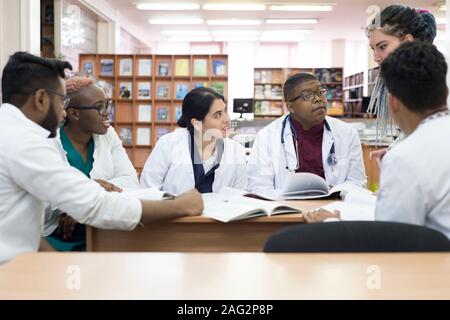 I giovani medici, razza mista. Essi siedono al tavolo, risolvere problemi medici. Le persone di sesso diverso, camici, abbigliamento medico, sono seduti ad un Foto Stock