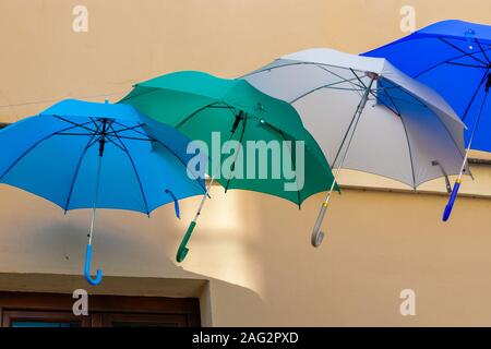 Ombrelli in diversi colori sullo sfondo di una parete su una soleggiata giornata estiva. L'azzurro, verde, grigio e blu ombrelloni appendere in aria contro Foto Stock