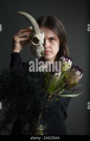 Ritratto di una giovane ragazza dai capelli scuri tenendo la metà di un teschio di capra davanti al suo volto e un mazzo di nero proteas e asparagi, doppiezza concept Foto Stock