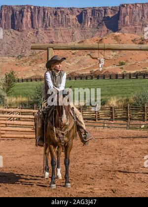 Un giovane di lavoro attraente cowgirl wrangler siede sul suo cavallo in un ranch vicino a Moab, Utah. Dietro di lei sono le scogliere di arenaria del canyon del Co Foto Stock