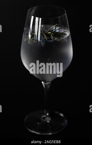 Primo piano di un bicchiere di vino con ghiaccio freddo il Gin Tonic con un rametto di rosmarino per il sapore, isolato prima di uno sfondo nero. Foto Stock