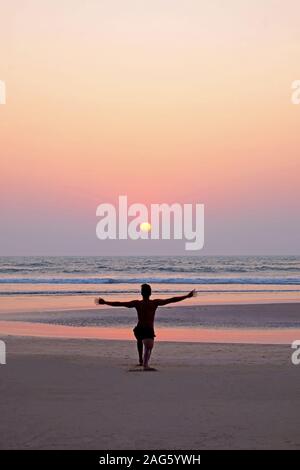 Un irriconoscibile uomo facendo un po' di yoga asana saluto al sole su di una vasta apertura vuota spiaggia sabbiosa, egli è inginocchiato con le braccia tese, egli è rivestito u Foto Stock