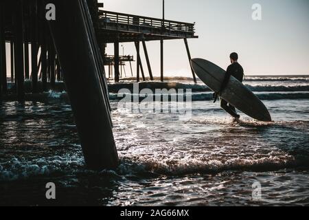 Surfboarder che trasporta una tavola da surf nell'oceano ondulato vicino a. molo in legno al tramonto Foto Stock