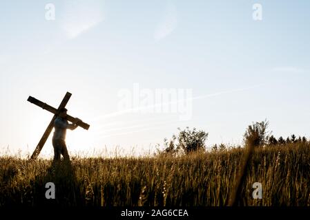 Il maschio che porta una mano fece una croce di legno in a. campo erboso con il sole che splende sullo sfondo Foto Stock