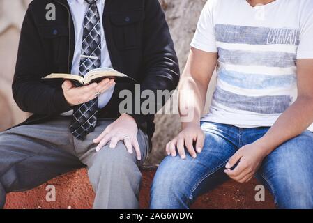 Colpo di primo piano di due maschi seduti mentre uno di loro legge la bibbia con sfondo sfocato Foto Stock