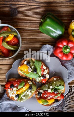 Grigliate le fette di pane condito con cotto rosso e verde e il peperone giallo, olive nere, rucola e formaggio Feta su sfondo di legno Foto Stock