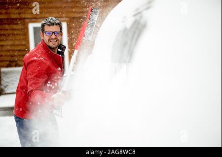 Uomo che indossa una giacca invernale rossa e pulisce la neve il finestrino della vettura Foto Stock