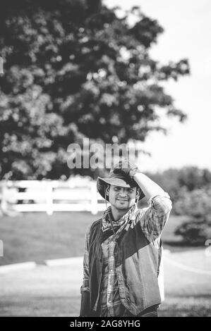 Immagine verticale in scala di grigi di un maschio che indossa un vestito da cowboy con la mano sul cappello Foto Stock