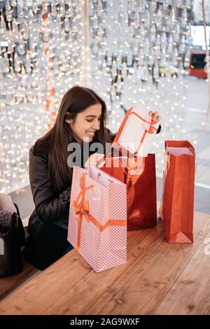 Giovane ragazza tenendo fuori avvolto regalo di Natale. Ragazza sorridente tirando regalo di Natale da una borsa regalo sul tavolo di legno al cafe con luci, con copia Foto Stock