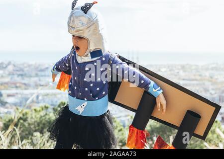 Funny bambino bambina gioca supereroe con costumi fatti in casa, cartone piano ali e astronauta casco battenti di fronte a un paesaggio urbano, l'immaginazione Foto Stock