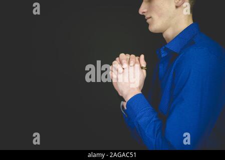 Uomo che indossa una t-shirt blu che prega con uno sfondo nero Foto Stock