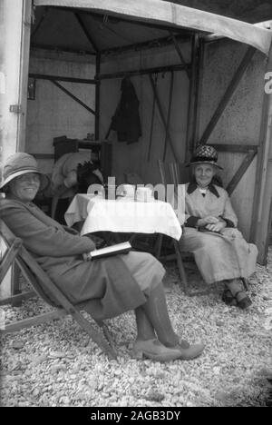 1930s, storico, due Signore nel vestito formale della giornata seduti in sdraio presso la loro capanna di mare, con una tazza di tè, Inghilterra, Regno Unito. Foto Stock