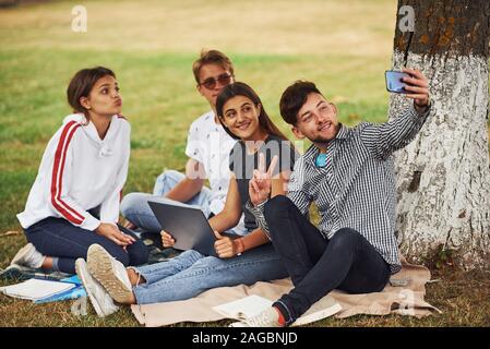 Seduta vicino alla struttura ad albero e rendendo selfie. Un gruppo di giovani studenti in abiti casual su erba verde di giorno Foto Stock