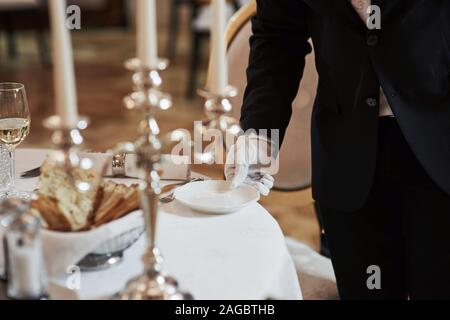 Trattiene la piastra bianca. Procedimento per la preparazione di luogo speciale per i visitatori. Cameriere in classiche opere di usura sul servering Foto Stock