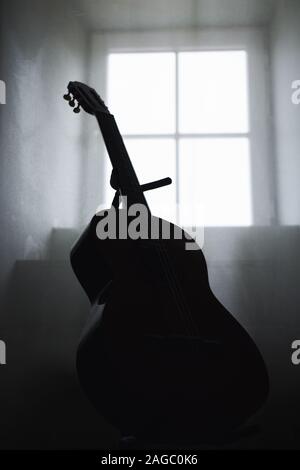 Immagine verticale in scala di grigi di una chitarra acustica all'interno di un buio camera con una finestra sullo sfondo Foto Stock