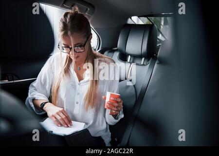 Mattina drink in mano. Smart imprenditrice si siede sul sedile posteriore della vettura di lusso con interni neri Foto Stock