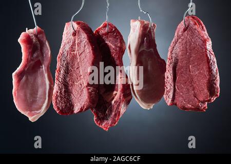 Un assortimento di carne cruda bistecche su ganci in metallo scuro su sfondo nero Foto Stock
