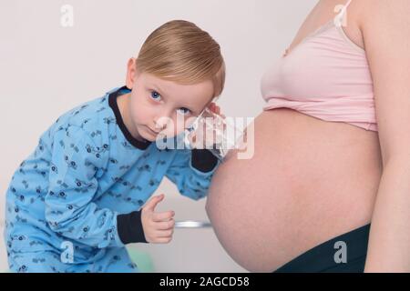 Ascolto di pancia. Curioso bello dark-eyed boy sensazione molto interessati mentre ascolto a donne in stato di gravidanza pancia di sua madre in stato di gravidanza. Foto Stock