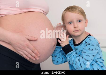 Ascolto di pancia. Curioso bello dark-eyed boy sensazione molto interessati mentre ascolto a donne in stato di gravidanza pancia di sua madre in stato di gravidanza. Foto Stock