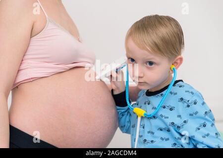 Ascolto di pancia. Curioso bello dark-eyed boy sensazione molto interessati mentre ascolto a donne in stato di gravidanza pancia di sua madre in stato di gravidanza. Foto Stock