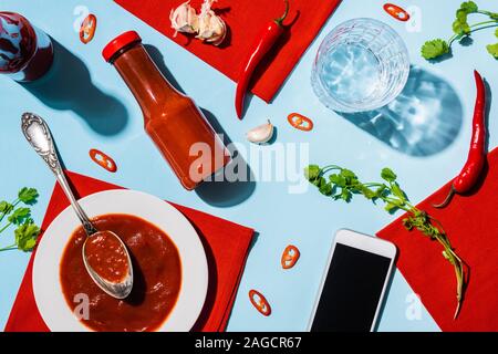 Vista superiore del ketchup fatti in casa con aglio e peperoncino accanto a smartphone con schermo vuoto sulla superficie blu Foto Stock