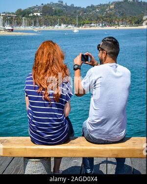 Una vista posteriore di un seduto con testa rossa giovane donna seduta a fianco di un giovane uomo che utilizza il suo telefono cellulare dotato di fotocamera digitale da un molo del porto di Santa Barbara, CA Foto Stock