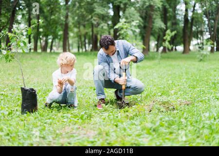 Padre di scavare con la pala vicino al figlio per piantare la piantina in posizione di parcheggio Foto Stock