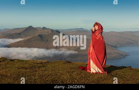 Una giovane donna dai capelli rossi in abiti vecchio stile con un mantello rosso rimane sul campo negli altopiani. Danimarca, isola di Faroe. Ho una versione del modello Foto Stock