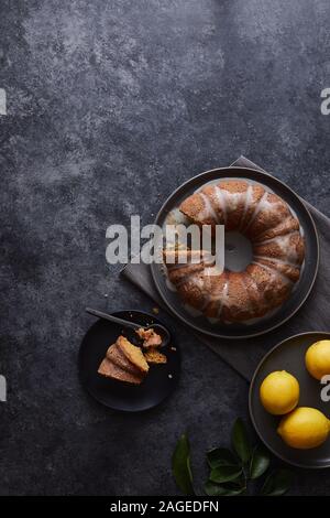 Colpo verticale di torta al limone coperto con salsa dolce vicino una piastra di limoni su una superficie nera Foto Stock