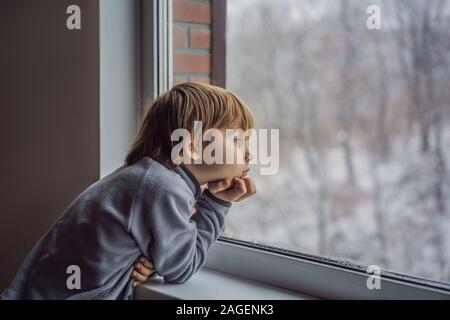 Felice adorabili kid ragazzo seduto vicino alla finestra e guardando al di fuori sulla neve il giorno di Natale o la mattina. Bambino sorridente affascinato con nevicata e grande Foto Stock