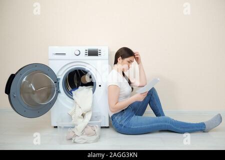 Sconvolto donna con biancheria sporca in scatola e manuale in mani si siede sul pavimento da una moderna macchina di lavaggio in camera Foto Stock