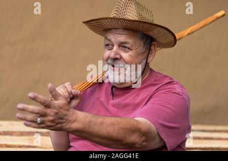 Senior barbuto uomo nel cappello di paglia seduto contro la parete di argilla, gesticulating con la mano sinistra mentre si tiene in vimini bastone in uno di destra sul dovrebbe Foto Stock
