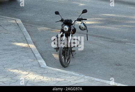 Un vecchio nero moto parcheggiata sul lato di una strada. Esso ha un casco Argento appeso al manubrio. Foto Stock