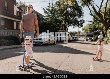 Padre e figlie giocando push scooter in quartiere Foto Stock