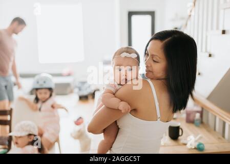 Madre con bambina in armi a casa, i membri della famiglia in background Foto Stock