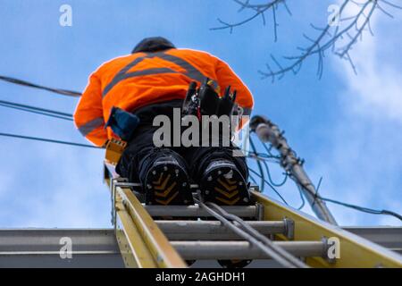 Un professionista telecoms installer è visto da sotto, l'installazione di cavi in fibra ottica dalla cima di una scala sul tetto di una abitazione di proprietà Foto Stock