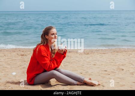 Bionda con una tazza di caffè si trova su una spiaggia di sabbia e guardando il mare Foto Stock