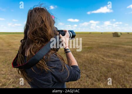 Giovane fotografo femmina con i capelli lunghi, visto da dietro e foto all'aperto verso l'orizzonte in un grande campo d'oro o di prato. Foto Stock