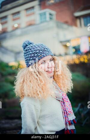 Ritratto di giovane sorridente curly donna bionda in bianco caldo maglione naturali e lavorati a maglia blu beanie hat con pompon guardando la fotocamera con il verde par Foto Stock