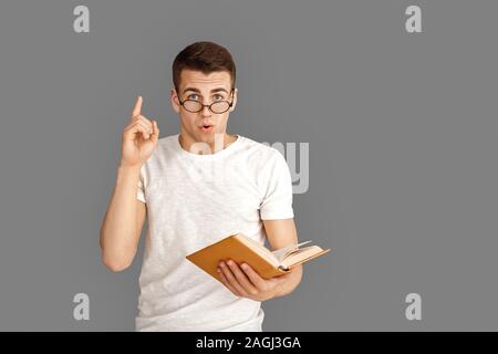 Freestyle. Giovane studente in occhiali in piedi isolato su grigio con libro rivolto verso l'alto idea Foto Stock