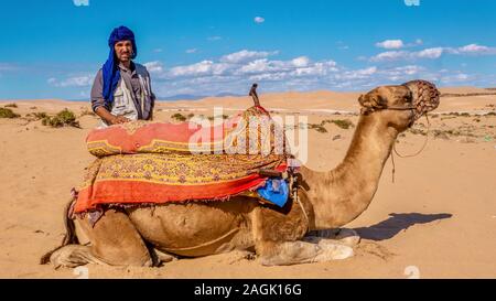 Agadir, Marocco - Ott. 21, 2015. Un uomo marocchino è la vendita di giri in cammello nel deserto per turisti. Il cammello è in ginocchio per un turista a monte. Foto Stock