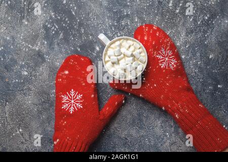 Natale e Anno Nuovo accogliente vacanza composizione con sciarpa, donna mani in muffole, tazze con bevanda calda e marshmallow sul cemento grigio Foto Stock