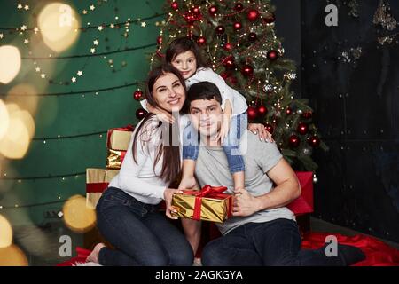 In posa per la foto. Bella famiglia siede vicino all'albero di Natale con confezioni regalo su inverno sera, godendo il tempo trascorrere insieme Foto Stock